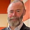 Headshot of Mark Goodchild, a smiling man with grey hair and a beard, wearing a collared shirt and black jacket, posed against a striped backdrop