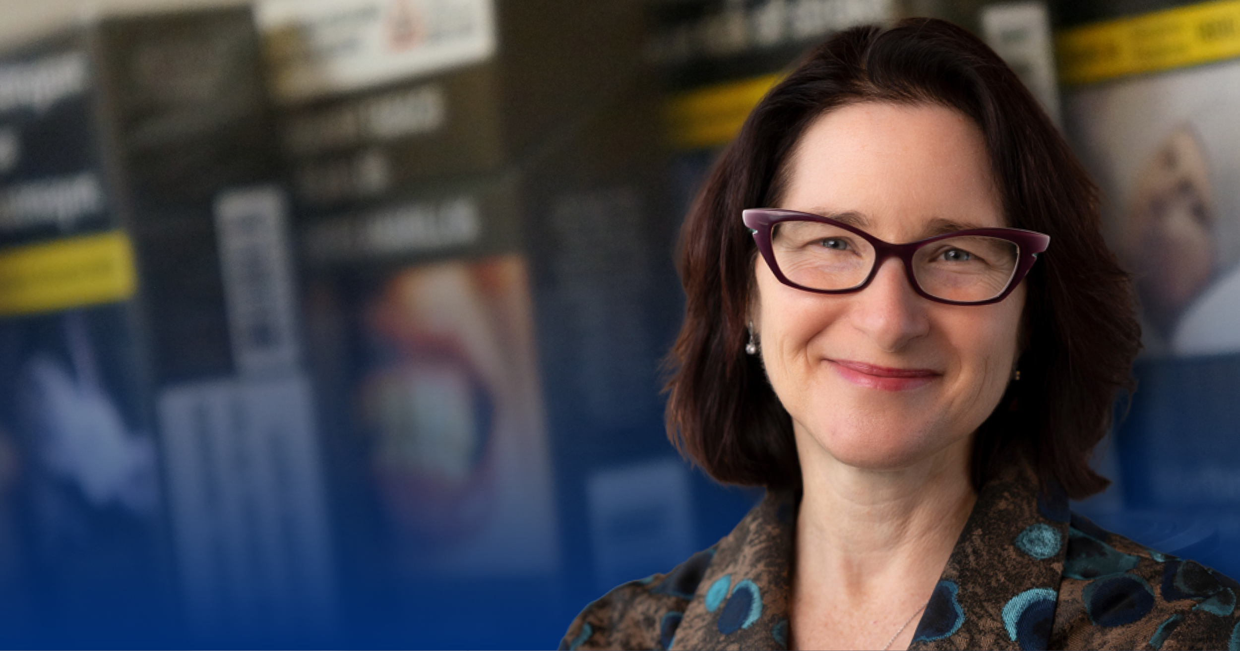 Headshot of Joanna Cohen, a smiling woman with wavy, brown, shoulder-length hair and a light complexion, wearing purple eyeglasses and a blouse with a brown and blue pattern, posed in front of a row of blurred cigarette packs with plain and standardized packaging