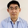 Headshot of Dr. Hiromasa Okayasu, a man with dark hair, eyeglasses, wearing a light blue collared shirt and posed against an off-white backdrop
