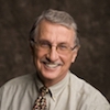 Headshot of Steve Tamplin, a smiling man with grey hair, a moustache, wearing a tan collared shirt with patterned necktie, posed against a dark backdrop