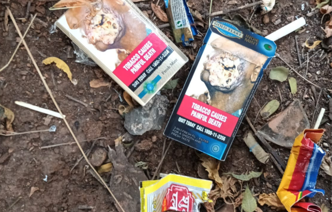 Assorted tobacco product litter with visible brand names and logos is strewn across the ground atop dirt twigs and leaves in a natural environment in India