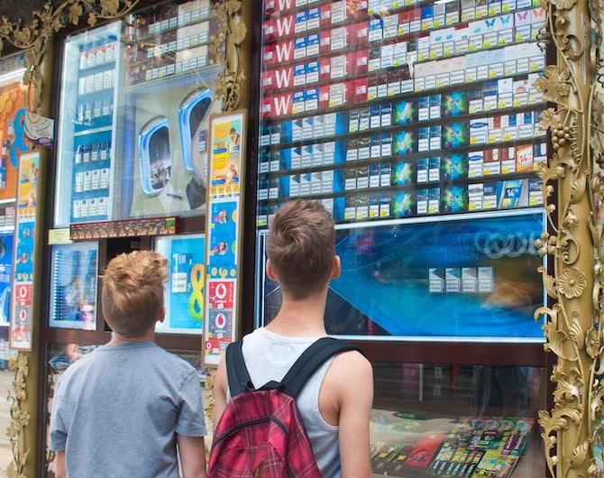 Two young boys standing outside a cigarette kiosk, facing a massive wall of cigarette advertising and products on display